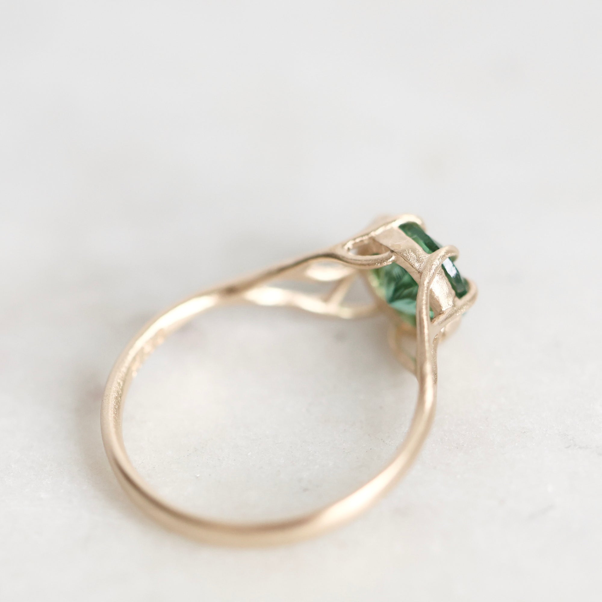 A delicate gold ring featuring a small green rectangular gemstone in a prong setting, resting on a light gray surface.