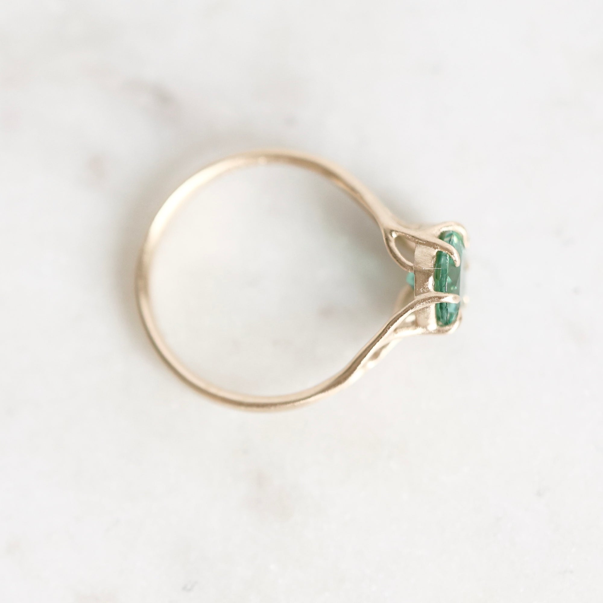 A gold ring with a unique twisted band and a small green gemstone cluster, photographed on a light marble surface.