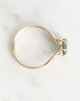 A gold ring with a unique twisted band and a small green gemstone cluster, photographed on a light marble surface.