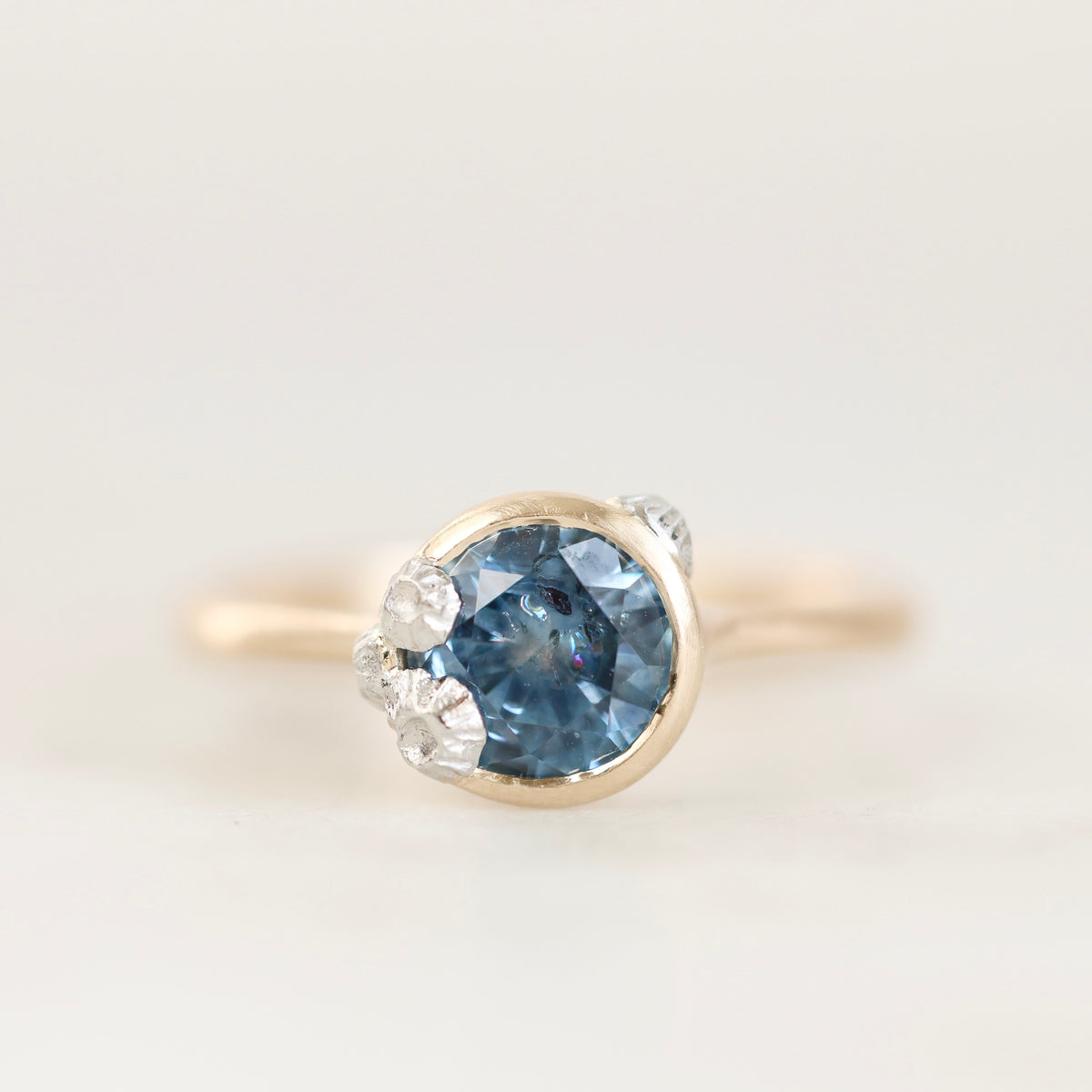 A yellow gold ring featuring a round-cut blue gemstone in a bezel setting, accented by small clear barnacle details on the shoulders, set against a soft white background.