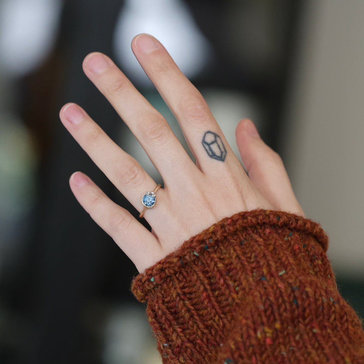 A close-up of a person's hand wearing a delicate gold ring with a round blue gemstone, set against a blurred background with a rust-colored knit sweater sleeve visible.