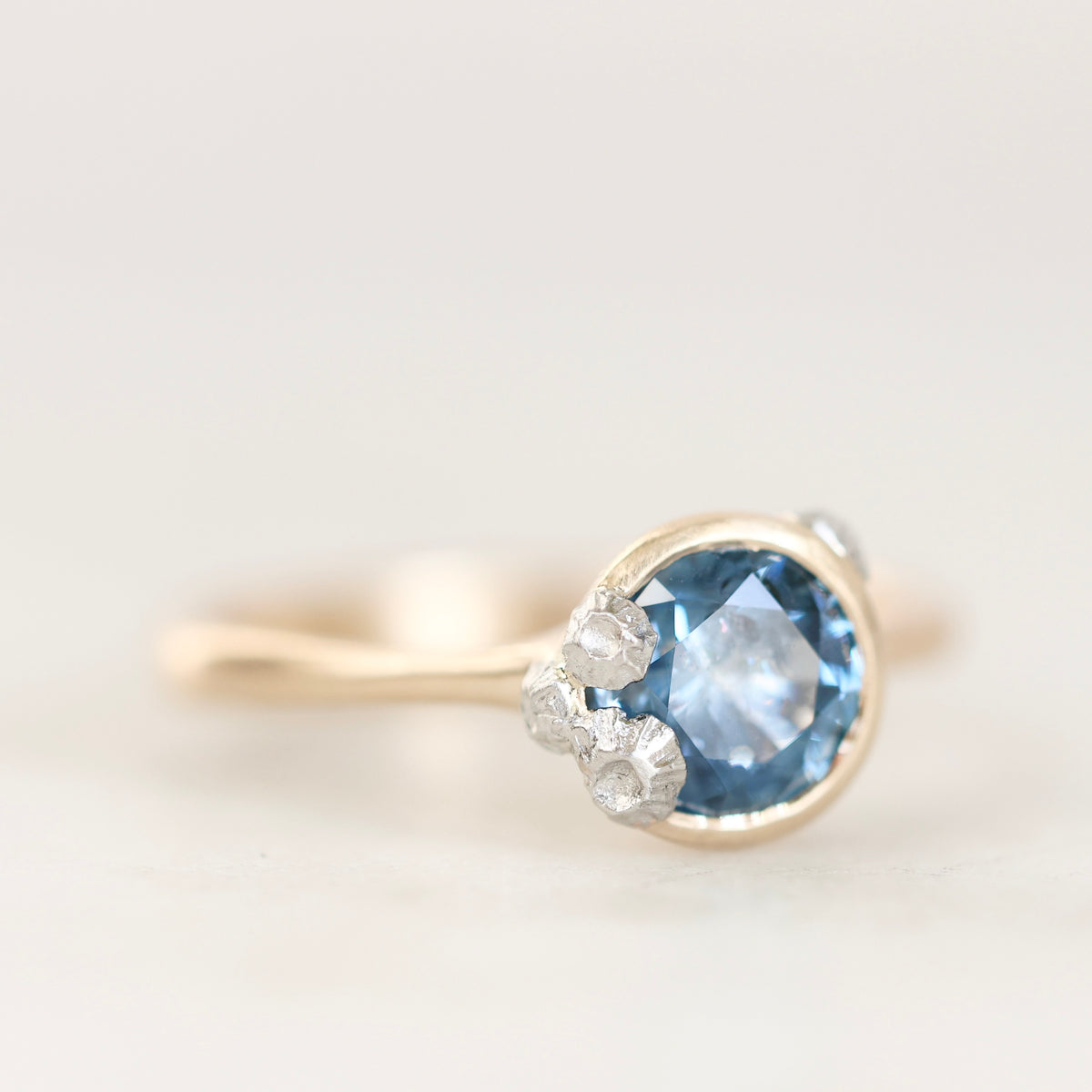 A close-up shot of a gold ring featuring a round-cut light blue gemstone accented by three small barnacle details, set against a soft, light off-white background.