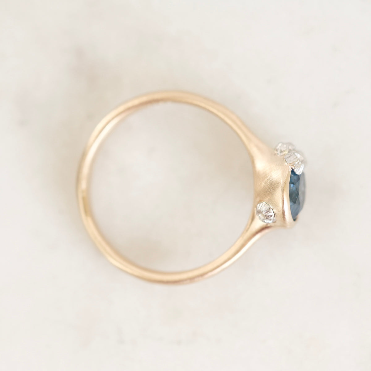 A close-up shot of a gold ring with a blue oval gemstone and two small barnacle details, resting on a light marble surface.