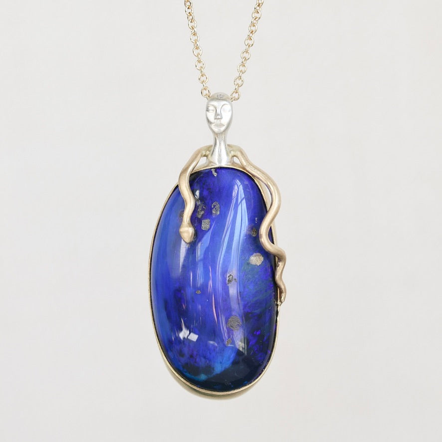 A pendant necklace featuring a large, polished blue opal stone with natural inclusions, set in a gold frame with a stylized face and snake-like details, on a light gray background.