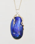 A pendant necklace featuring a large, polished blue opal stone with natural inclusions, set in a gold frame with a stylized face and snake-like details, on a light gray background.