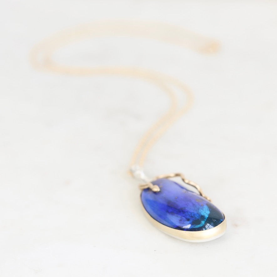 A teardrop-shaped pendant necklace featuring a vibrant blue iridescent stone in a gold bezel setting, suspended from a delicate gold chain on a light marble background.