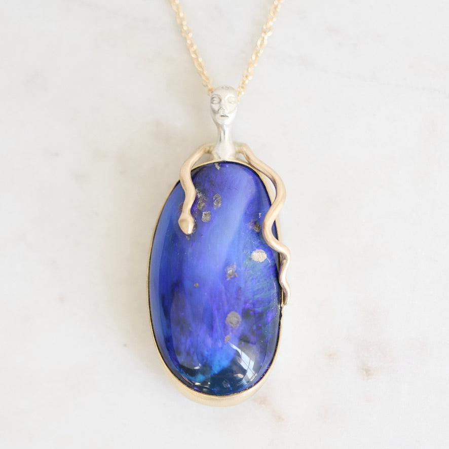 An oval pendant necklace featuring a deep blue opal stone with gold flecks, set in a silver-toned metal frame with a snake-like bail and a delicate gold chain, displayed on a light marble surface.