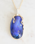 An oval pendant necklace featuring a deep blue opal stone with gold flecks, set in a silver-toned metal frame with a snake-like bail and a delicate gold chain, displayed on a light marble surface.