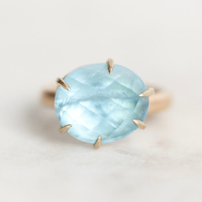 An oval-cut light blue gemstone ring in a gold prong setting, photographed on a clean, light off-white background.