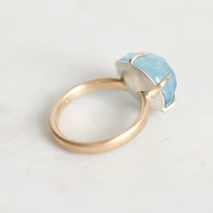 A minimalist ring featuring a polished gold band and a bezel-set light blue stone, photographed on a clean, light off-white surface.