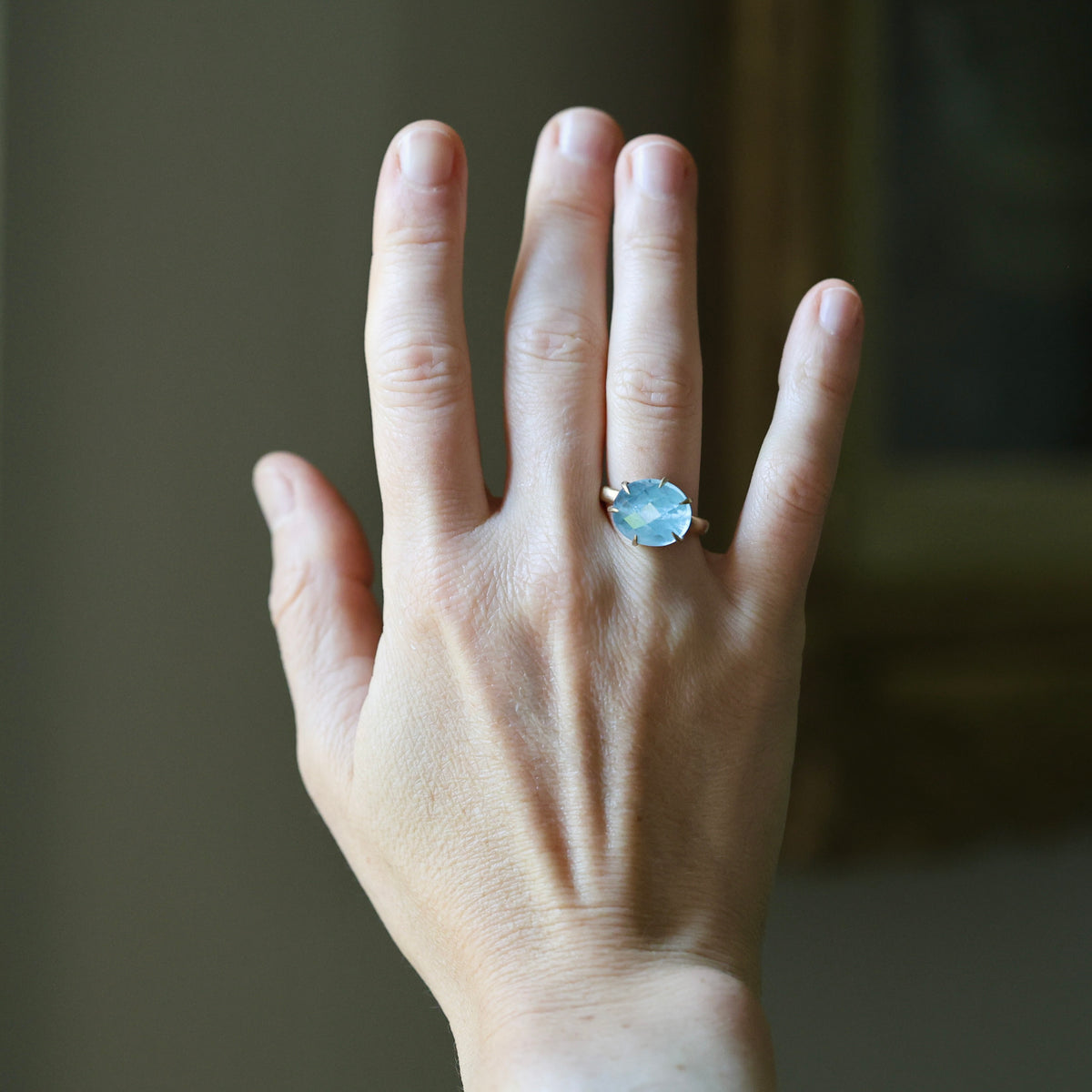 A close-up of a person's hand wearing a silver ring with a round light blue gemstone, set against a softly blurred indoor background.