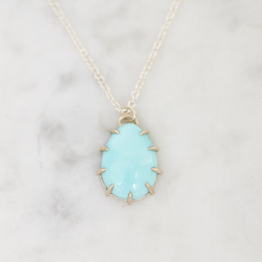 A silver necklace with a teardrop-shaped light blue gemstone pendant, displayed on a white marble surface.