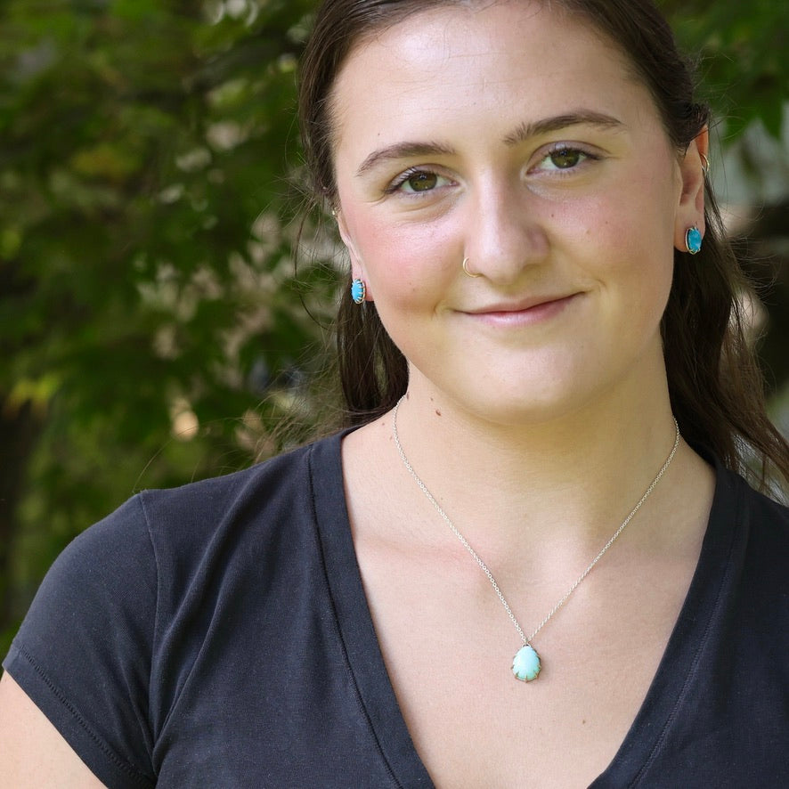 A woman with brown hair tied back, wearing a silver necklace with a light blue pendant and matching light blue stud earrings, photographed outdoors against a blurred green foliage background.
