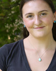 A woman with brown hair tied back, wearing a silver necklace with a light blue pendant and matching light blue stud earrings, photographed outdoors against a blurred green foliage background.