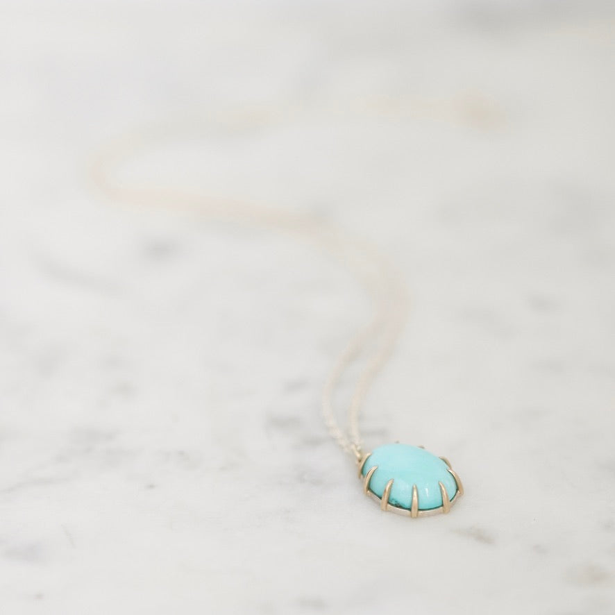 A delicate gold chain necklace featuring an oval pendant with a light turquoise blue center and gold prongs, resting on a white marble surface.