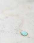 A delicate gold chain necklace featuring an oval pendant with a light turquoise blue center and gold prongs, resting on a white marble surface.