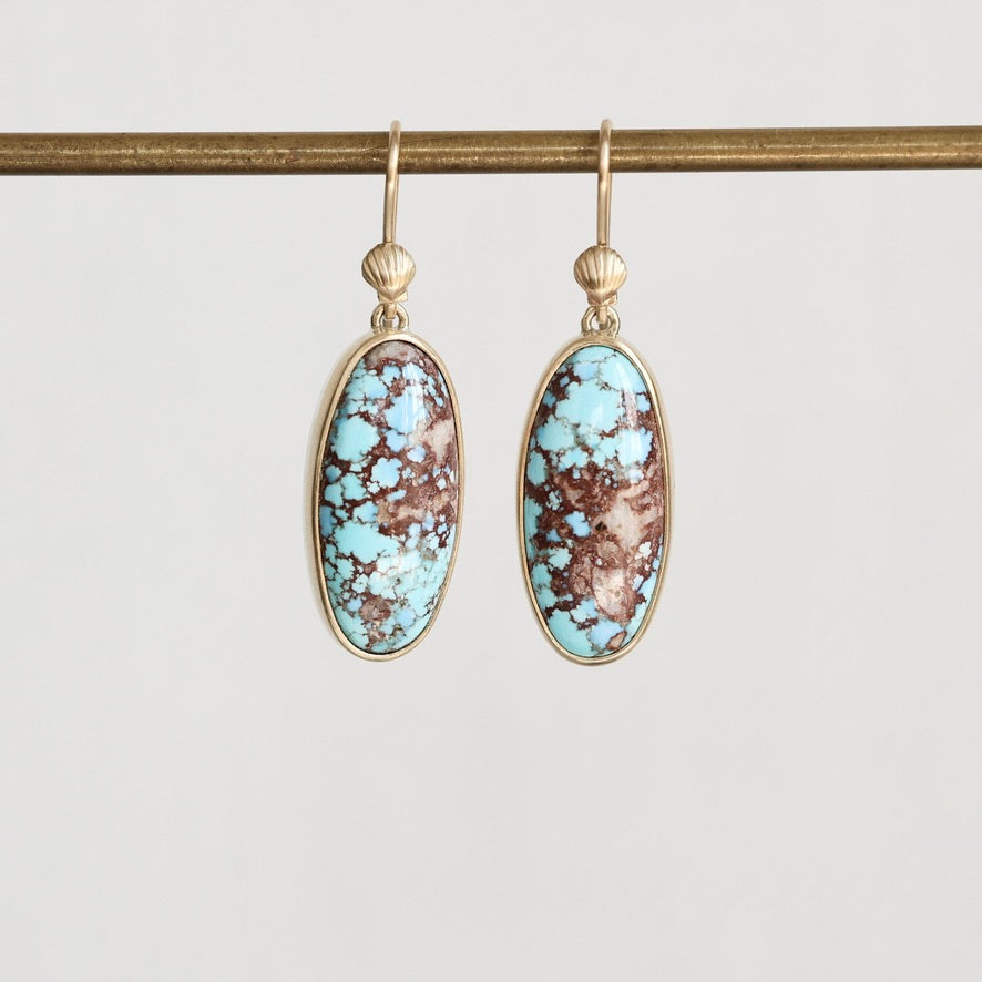 A pair of gold drop earrings with oval turquoise and brown marbled stone, hanging from a gold rod against a light gray background.