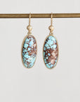A pair of gold drop earrings with oval turquoise and brown marbled stone, hanging from a gold rod against a light gray background.