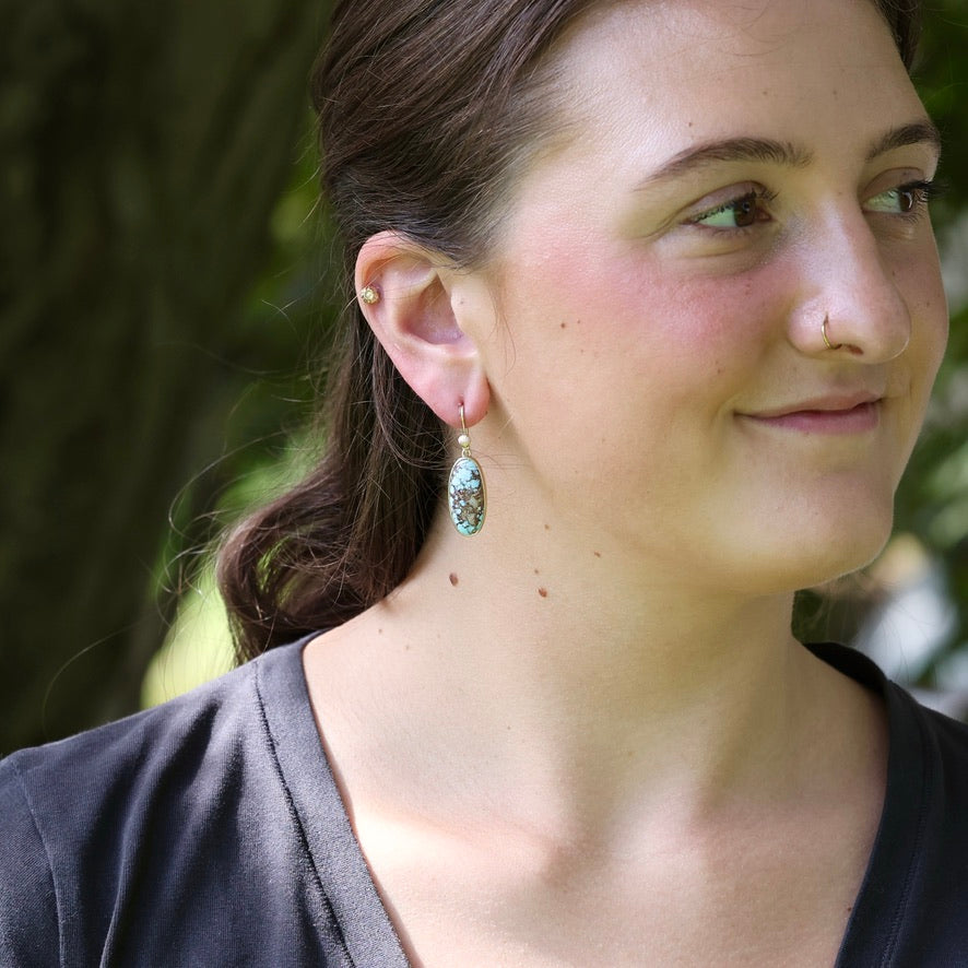 A woman wearing a dangling teardrop earring with a textured, glittery light blue-green stone in a gold-toned setting, photographed outdoors against a blurred green background.