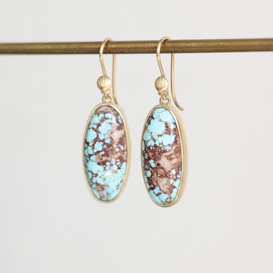 A pair of gold drop earrings with oval turquoise and brown marbled stone inlays, hanging from a gold rod against a light gray background.