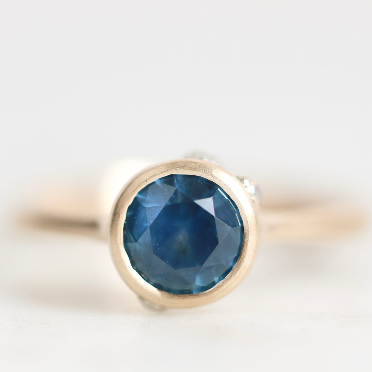 A close-up shot of a gold ring with a round-cut blue gemstone in a bezel setting, photographed against a soft, light gray background.