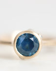 A close-up shot of a gold ring with a round-cut blue gemstone in a bezel setting, photographed against a soft, light gray background.