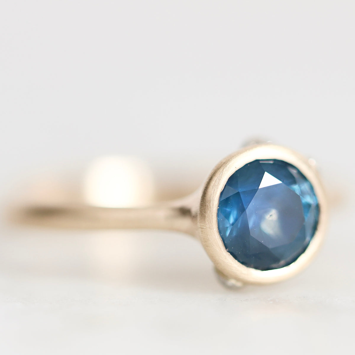 A gold ring with a round faceted blue gemstone, photographed in a close-up macro shot against a soft, light gray background.