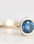 A gold ring with a round faceted blue gemstone, photographed in a close-up macro shot against a soft, light gray background.
