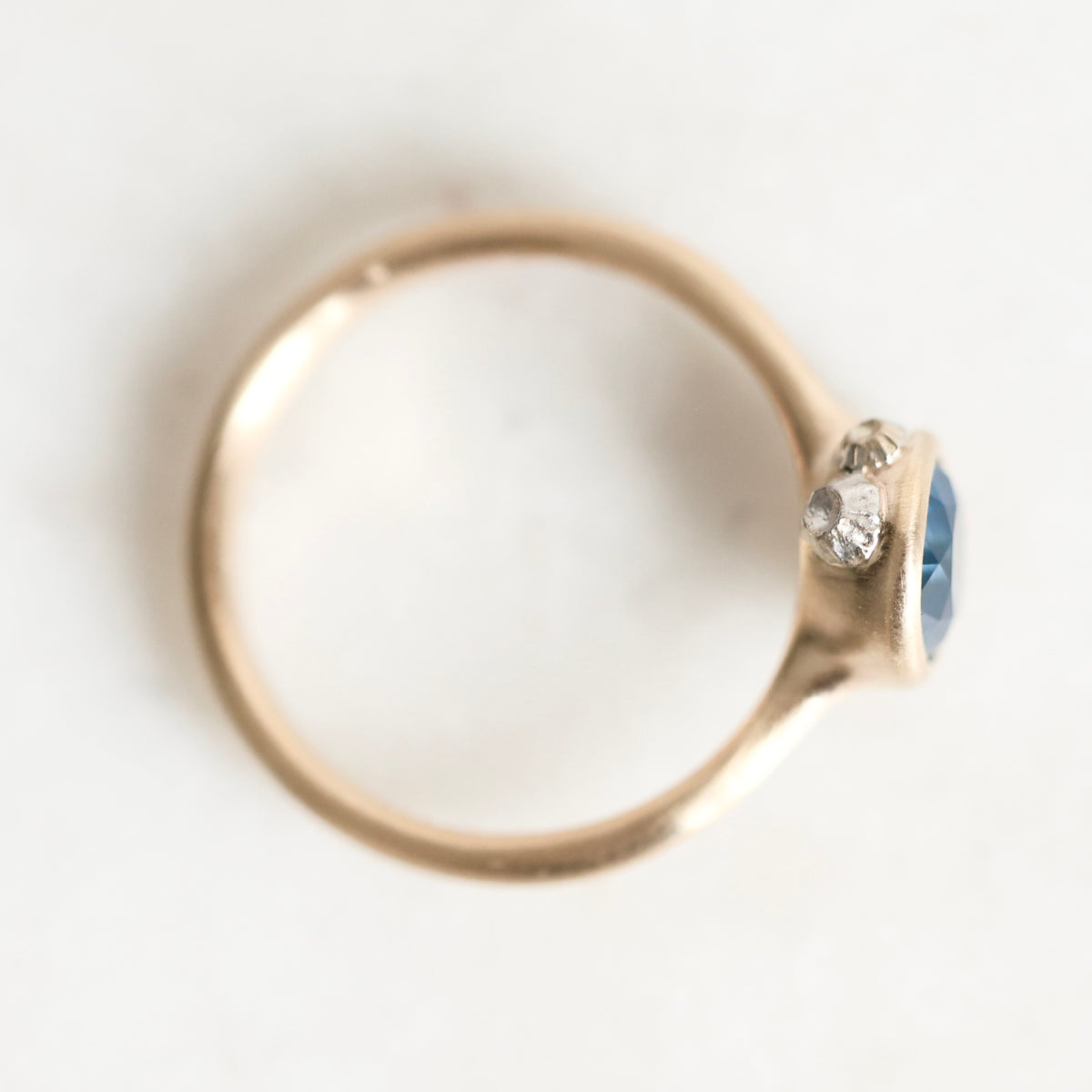 A close-up shot of a gold ring with a round blue gemstone and small clear accent stones, photographed on a plain white background.