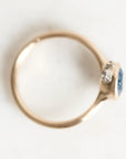 A close-up shot of a gold ring with a round blue gemstone and small clear accent stones, photographed on a plain white background.