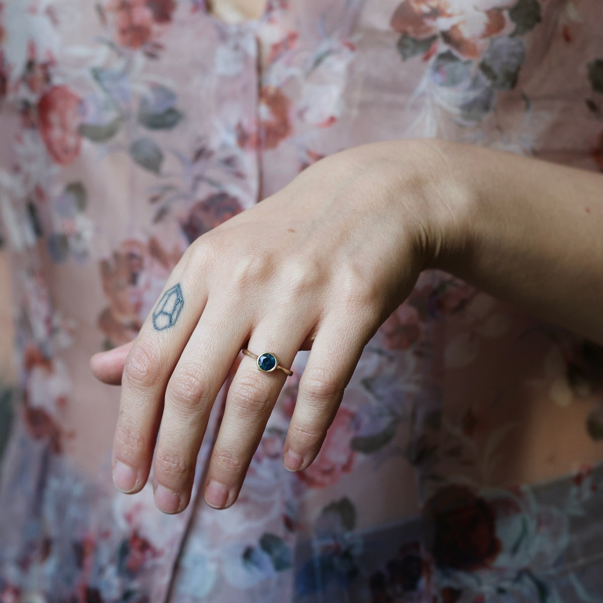 Womans hand with tattoo casually posed with a bezel set blue stone ring against a floral background. 