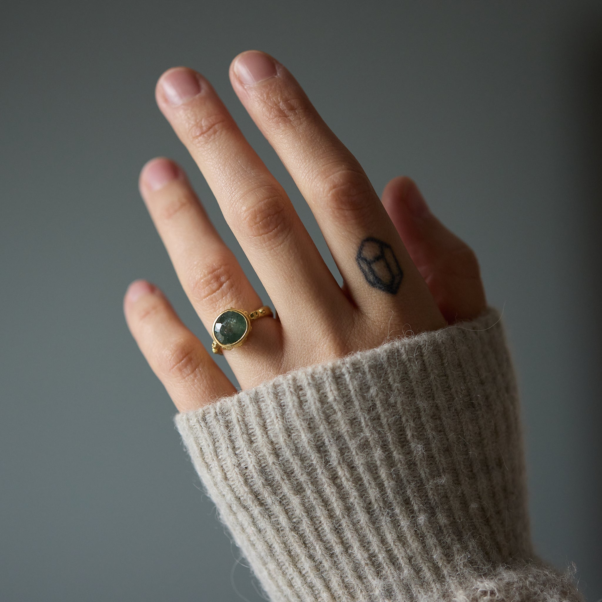 Hand wearing a gold ring with a green Montana sapphire, against a neutral background