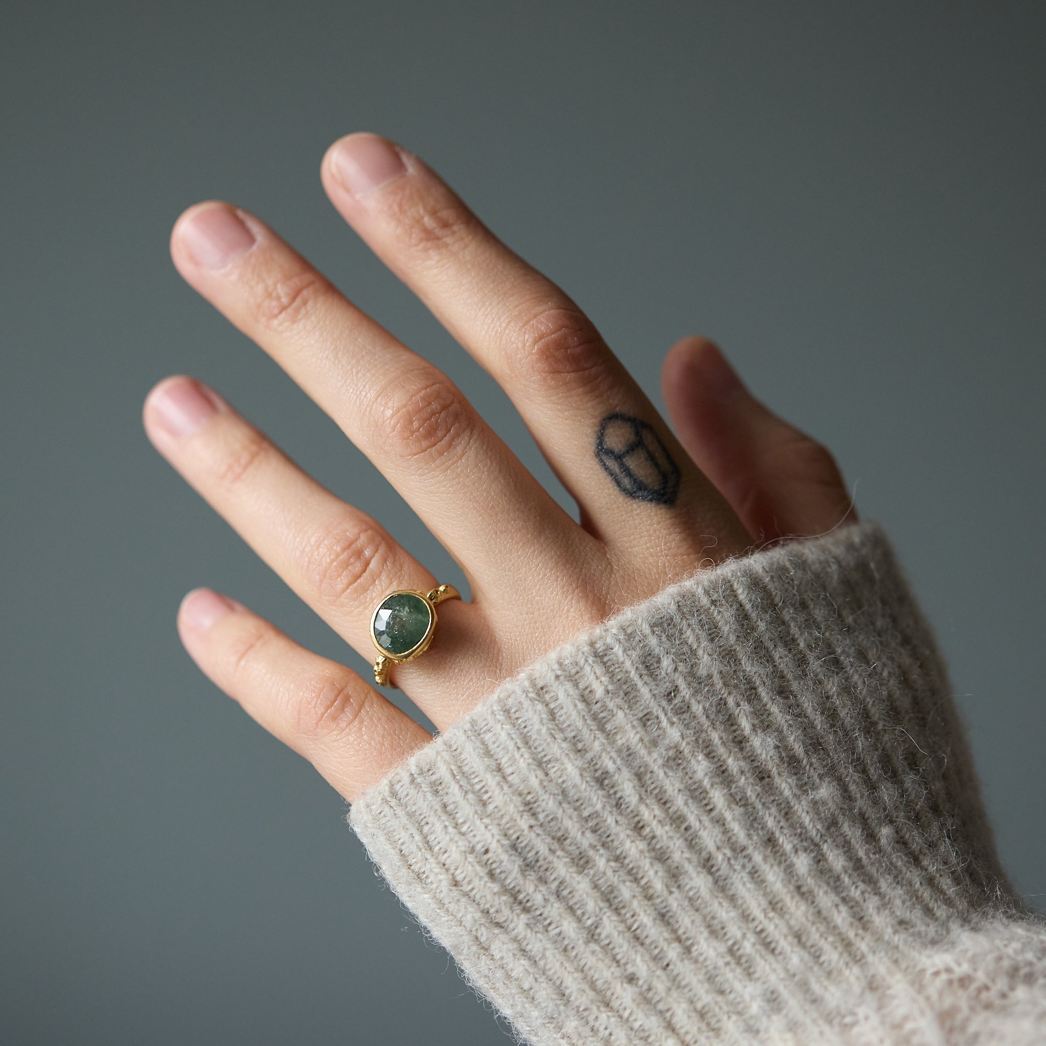 Hand wearing a gold ring with a green Montana Sapphire against a gray background