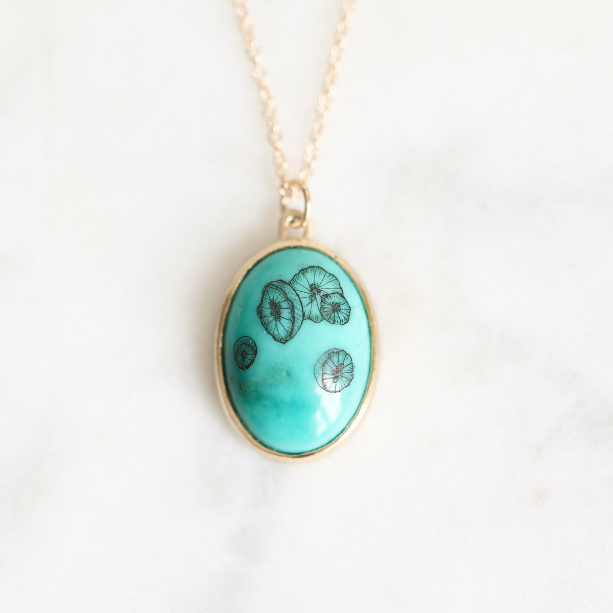 An oval turquoise pendant necklace with black mushroom illustrations, set in a gold-tone bezel on a delicate gold chain, displayed on a light marble surface.