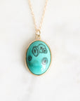 An oval turquoise pendant necklace with black mushroom illustrations, set in a gold-tone bezel on a delicate gold chain, displayed on a light marble surface.