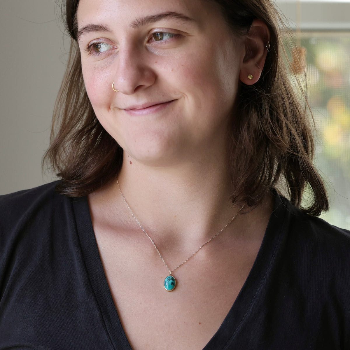 A woman with brown hair wearing a delicate gold chain necklace with a small oval turquoise pendant, paired with a black V-neck top.