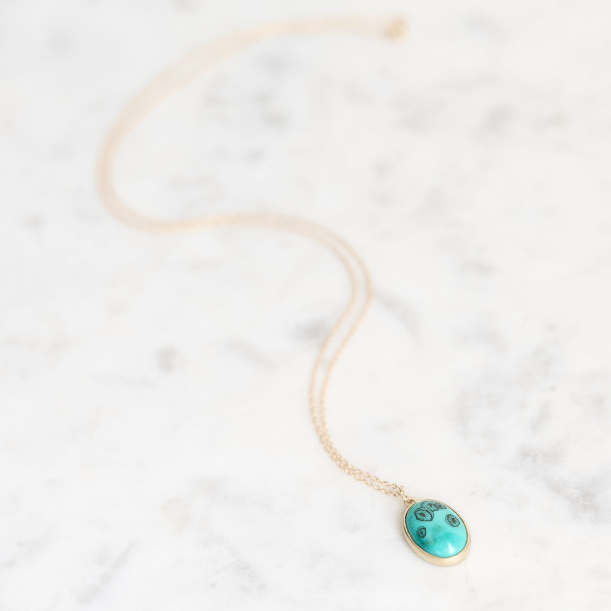 A delicate gold chain necklace with an oval turquoise pendant featuring black inlay details, resting on a white marble surface.