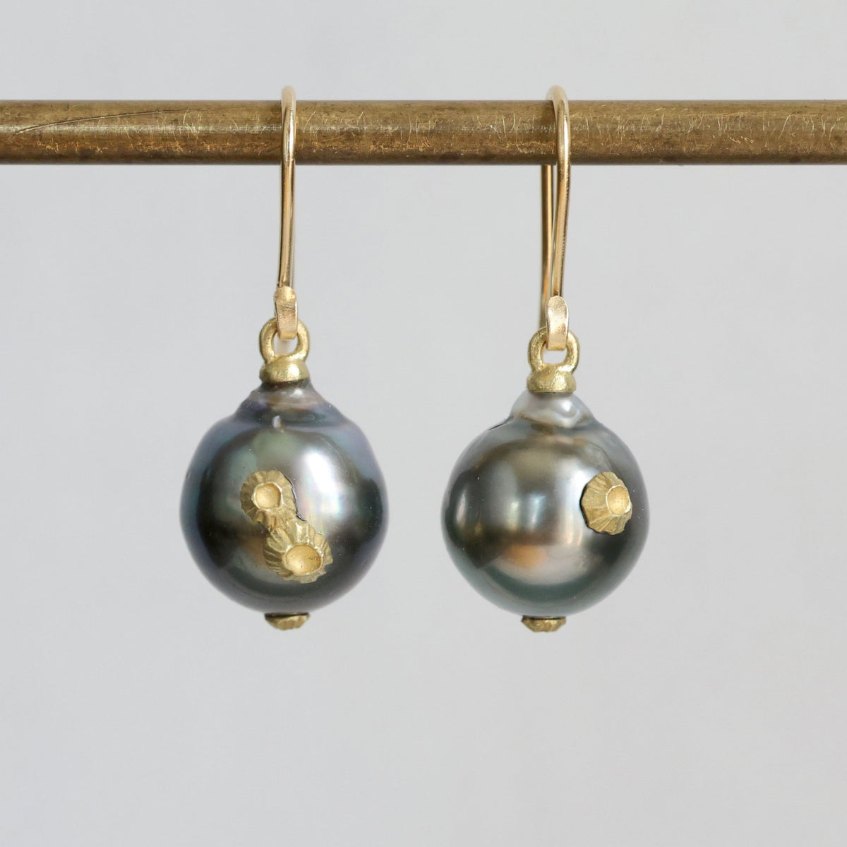 A pair of gold hook earrings with dark gray baroque pearls featuring gold inclusions, hanging from a gold rod against a light gray background.