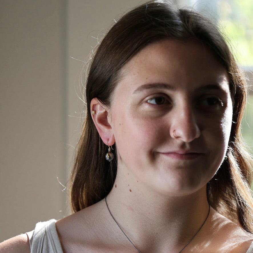 A close-up portrait of a young woman with brown hair, wearing a silver-toned dangle earring with a pearl-like bead and a delicate necklace, set against a softly blurred indoor background.