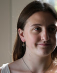 A close-up portrait of a young woman with brown hair, wearing a silver-toned dangle earring with a pearl-like bead and a delicate necklace, set against a softly blurred indoor background.