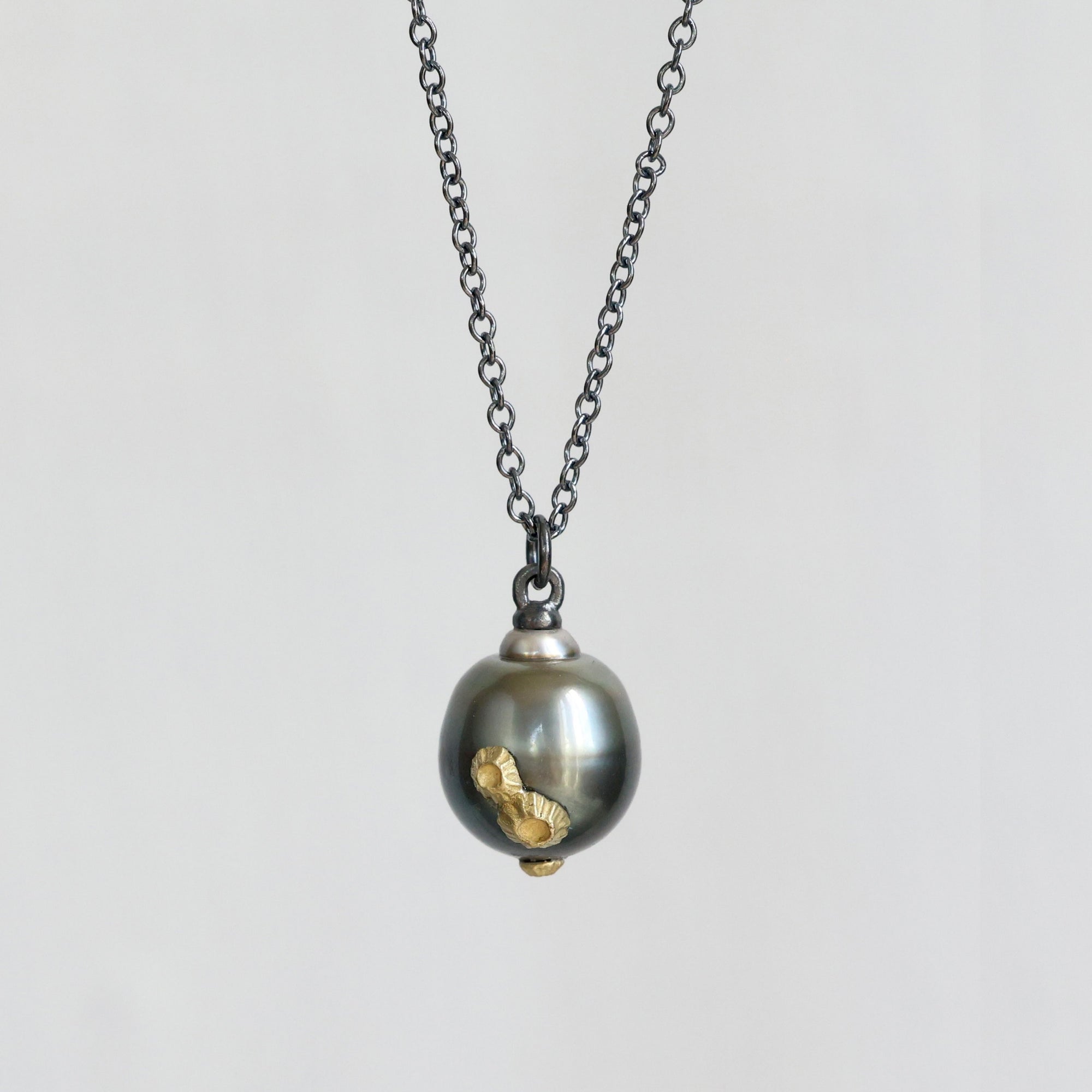 A dark gunmetal chain necklace with a pendant featuring a large dark gray pearl and a small gold-colored bird charm, set against a plain light gray background.