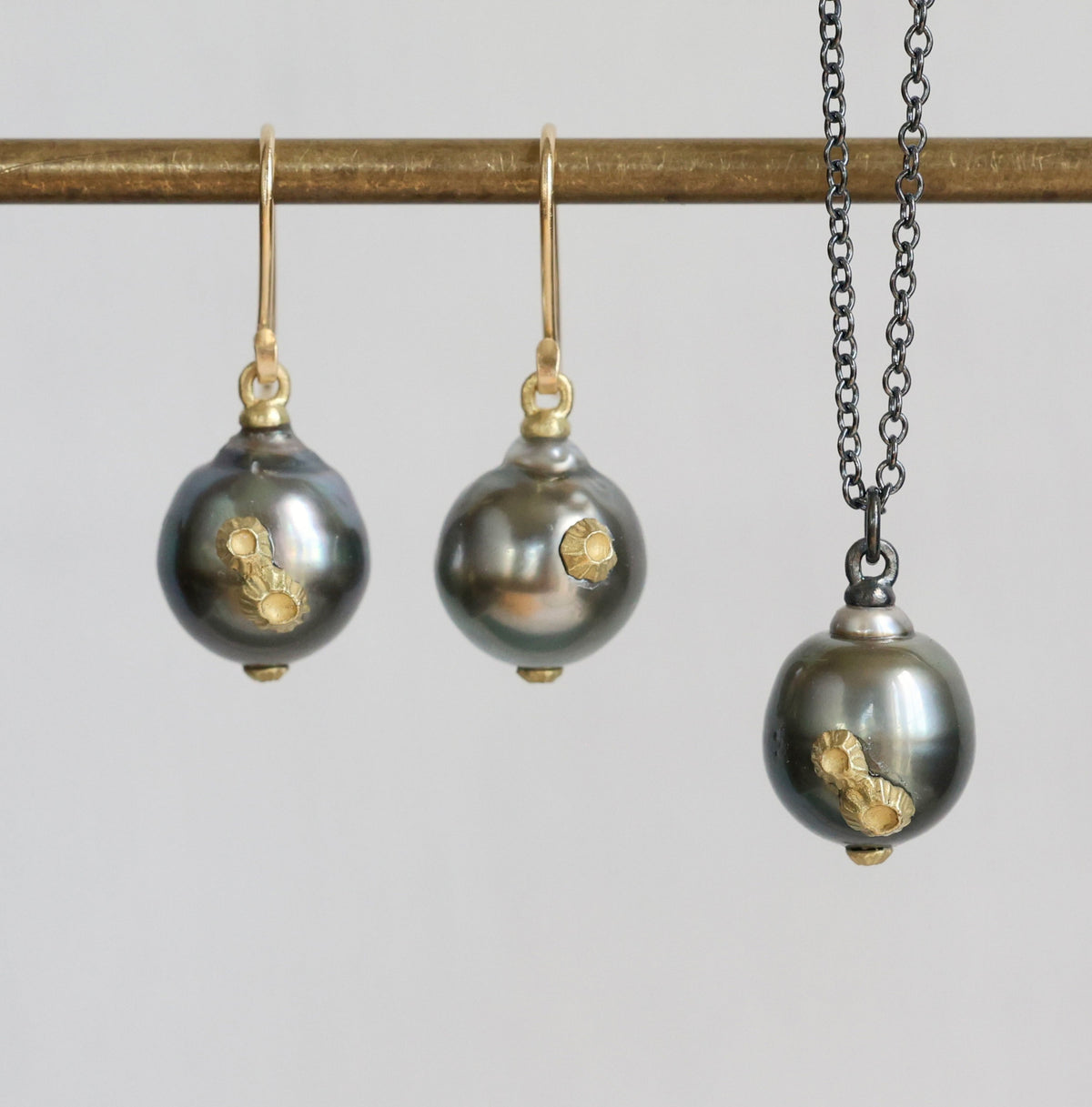 A jewelry set featuring a necklace and matching drop earrings with dark gray baroque pearls accented by gold leaf, displayed on a gold-colored rod against a light gray background.