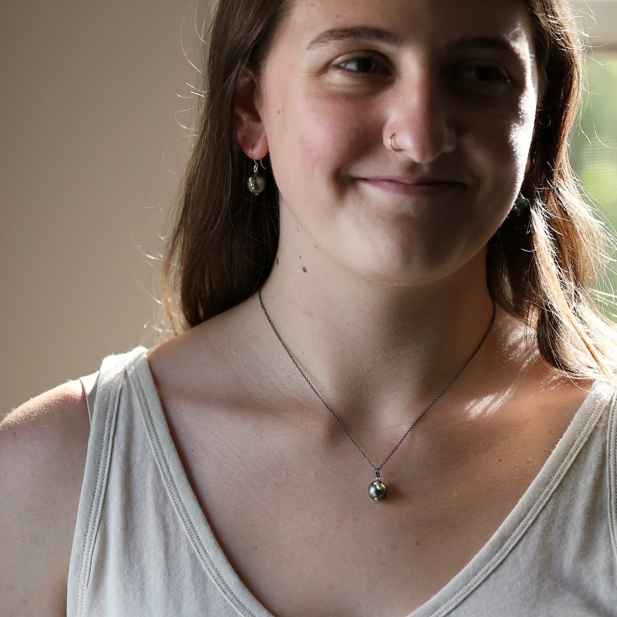 A close-up lifestyle shot of a woman wearing a delicate silver pendant necklace and matching drop earrings. The necklace features a small, polished silver bead pendant on a fine silver chain.