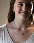 A close-up lifestyle shot of a woman wearing a delicate silver pendant necklace and matching drop earrings. The necklace features a small, polished silver bead pendant on a fine silver chain.