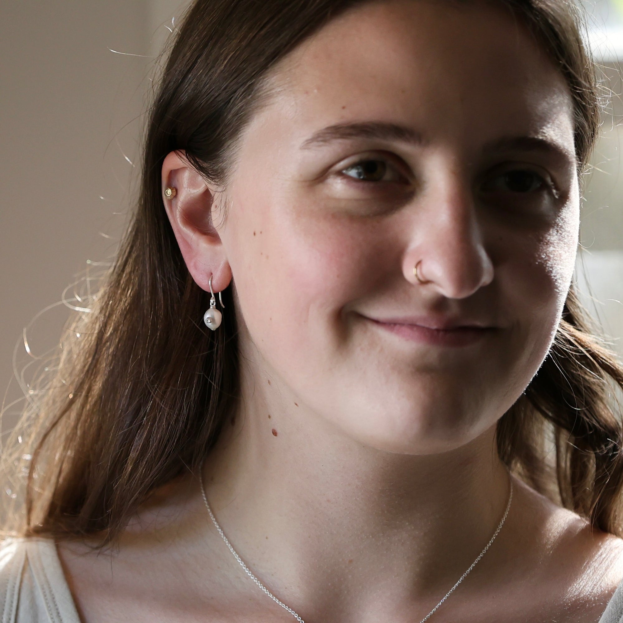 A close-up portrait of a woman wearing a delicate silver hoop earring with a small white bead drop, shown in warm indoor lighting.