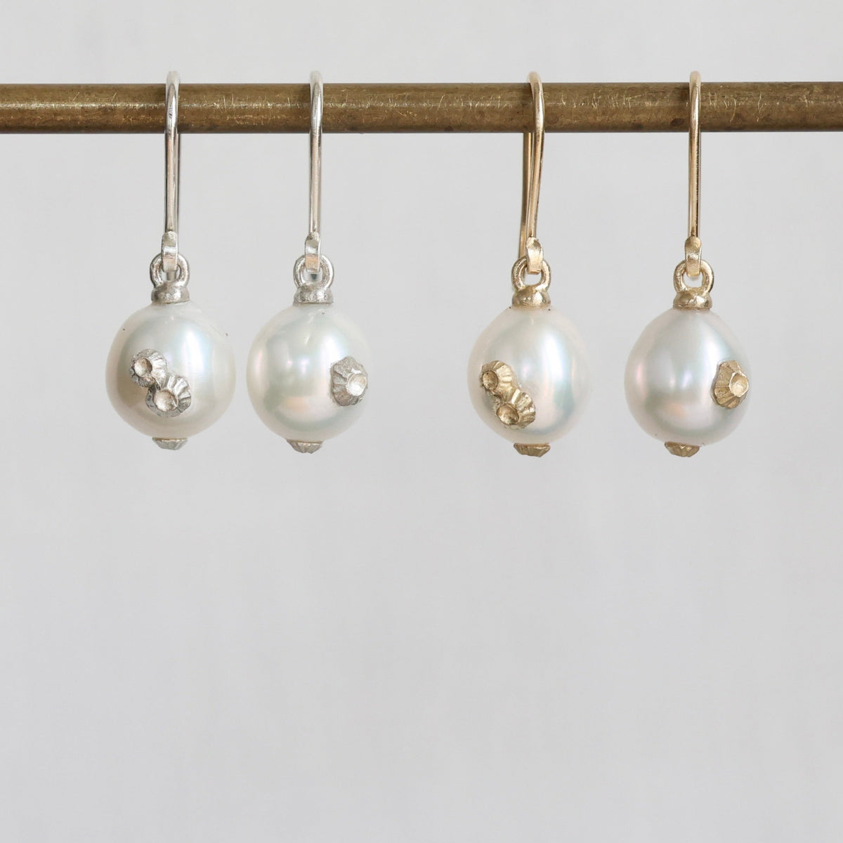 Four pearl drop earrings hanging from a gold-toned rod against a light gray background. Two earrings have white freshwater pearls with small silver floral accents, and two have white pearls with small gold floral accents.