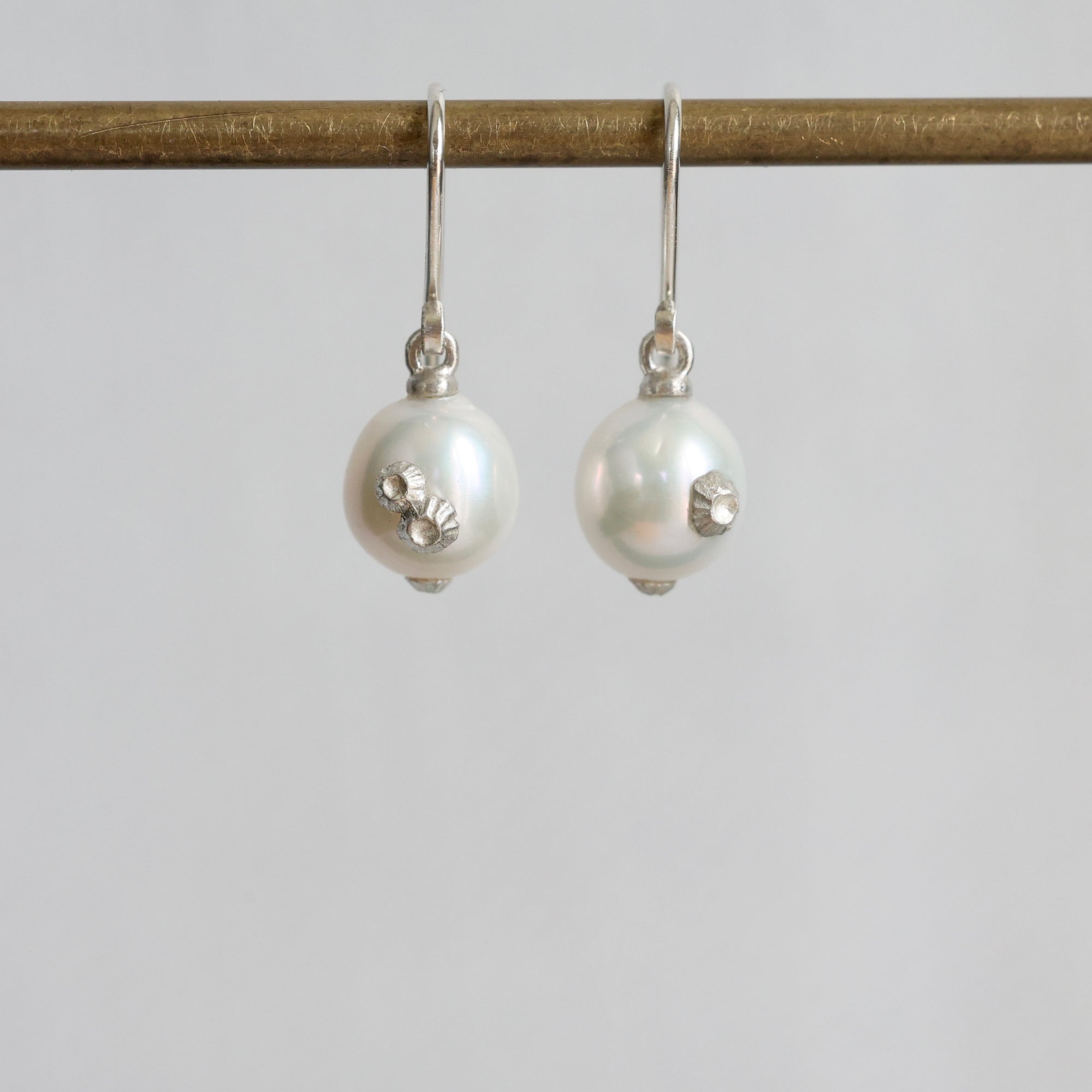 A pair of silver drop earrings with large white pearl beads and small silver floral embellishments, hanging from a gold-toned rod against a light gray background.