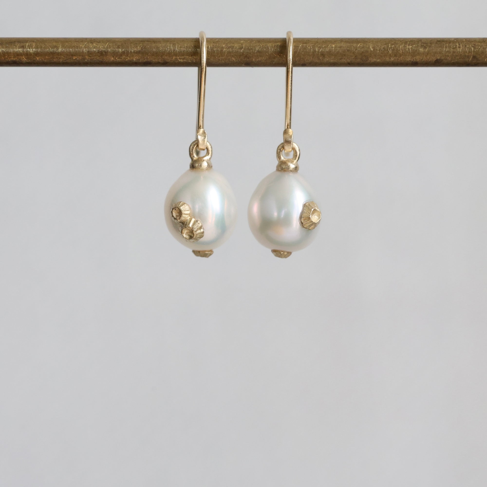 A pair of gold drop earrings with white pearl beads and small gold floral accents, hanging from a gold rod against a light gray background.
