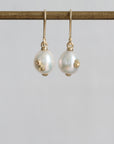 A pair of gold drop earrings with white pearl beads and small gold floral accents, hanging from a gold rod against a light gray background.