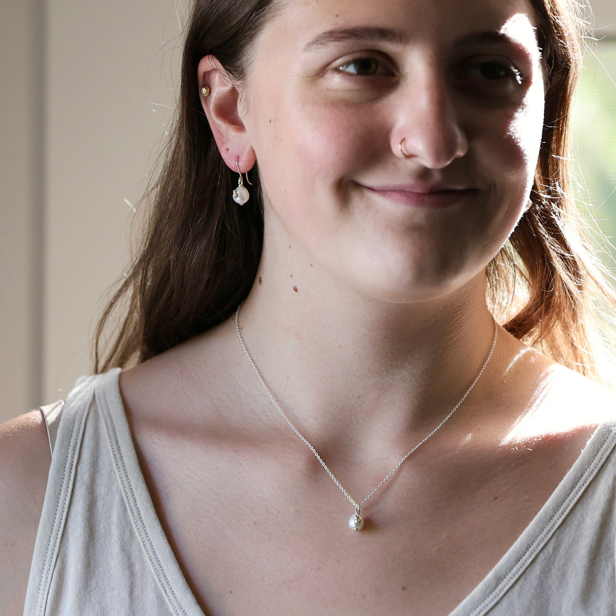 A close-up lifestyle shot of a woman wearing a delicate silver chain necklace with a single white pearl pendant, along with matching stud earrings.